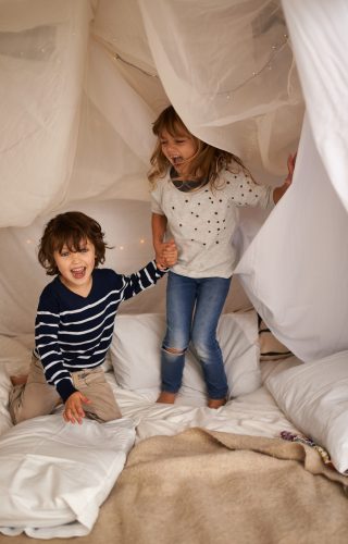 Shot of two adorable siblings jumping on the mattress underneath their fort